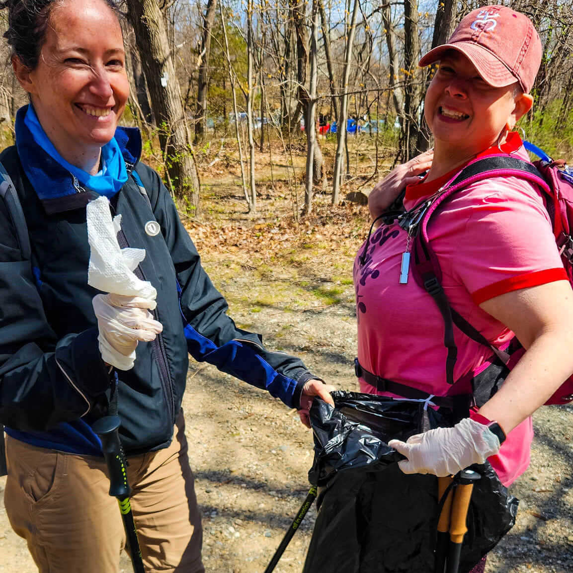 Two women on a trail, one holding hiking poles and the other with a trash bag, in a natural setting.