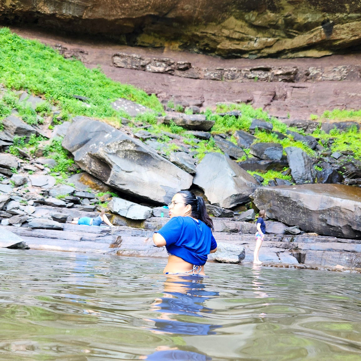 Person in a blue shirt wading in a natural pool with rocky and grassy banks.