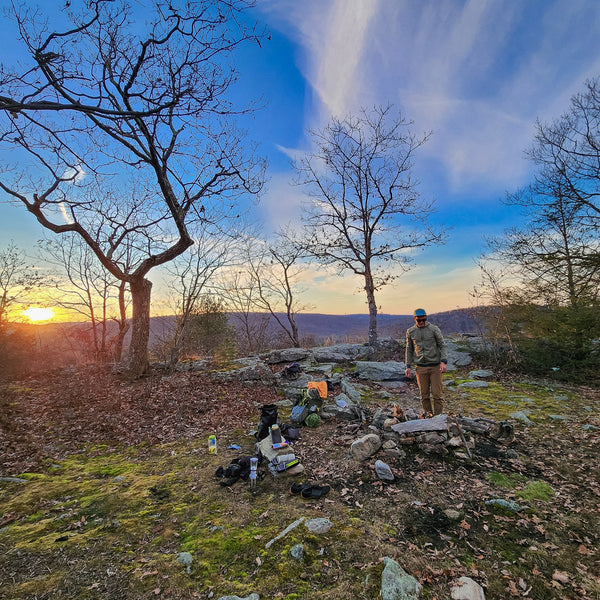 Person standing in a forest clearing with a campfire and camping gear at sunset.