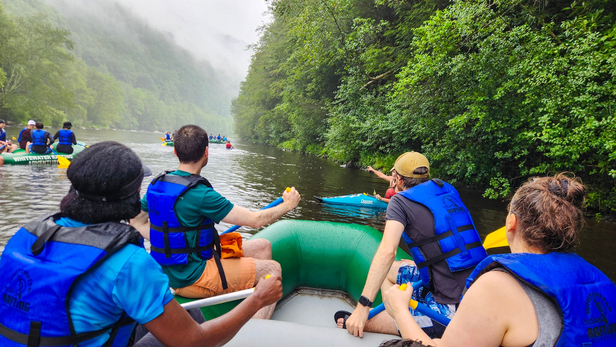 Group of people on a river rafting adventure with trees and foggy mountains in the background.