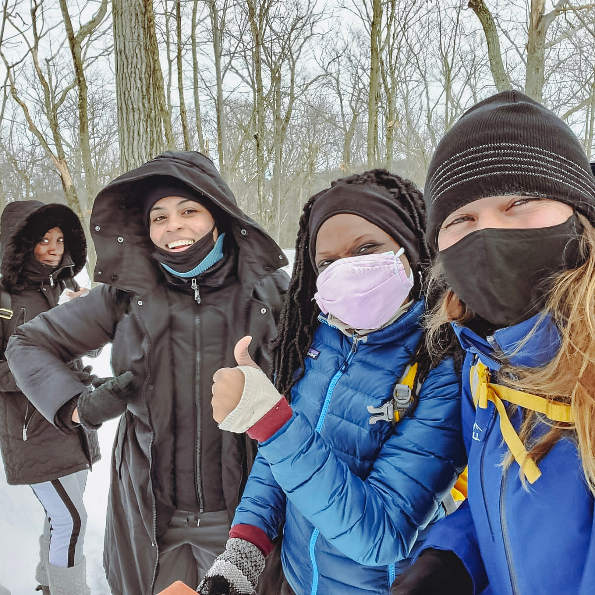 Four people in winter clothing and masks standing in a snowy forest.