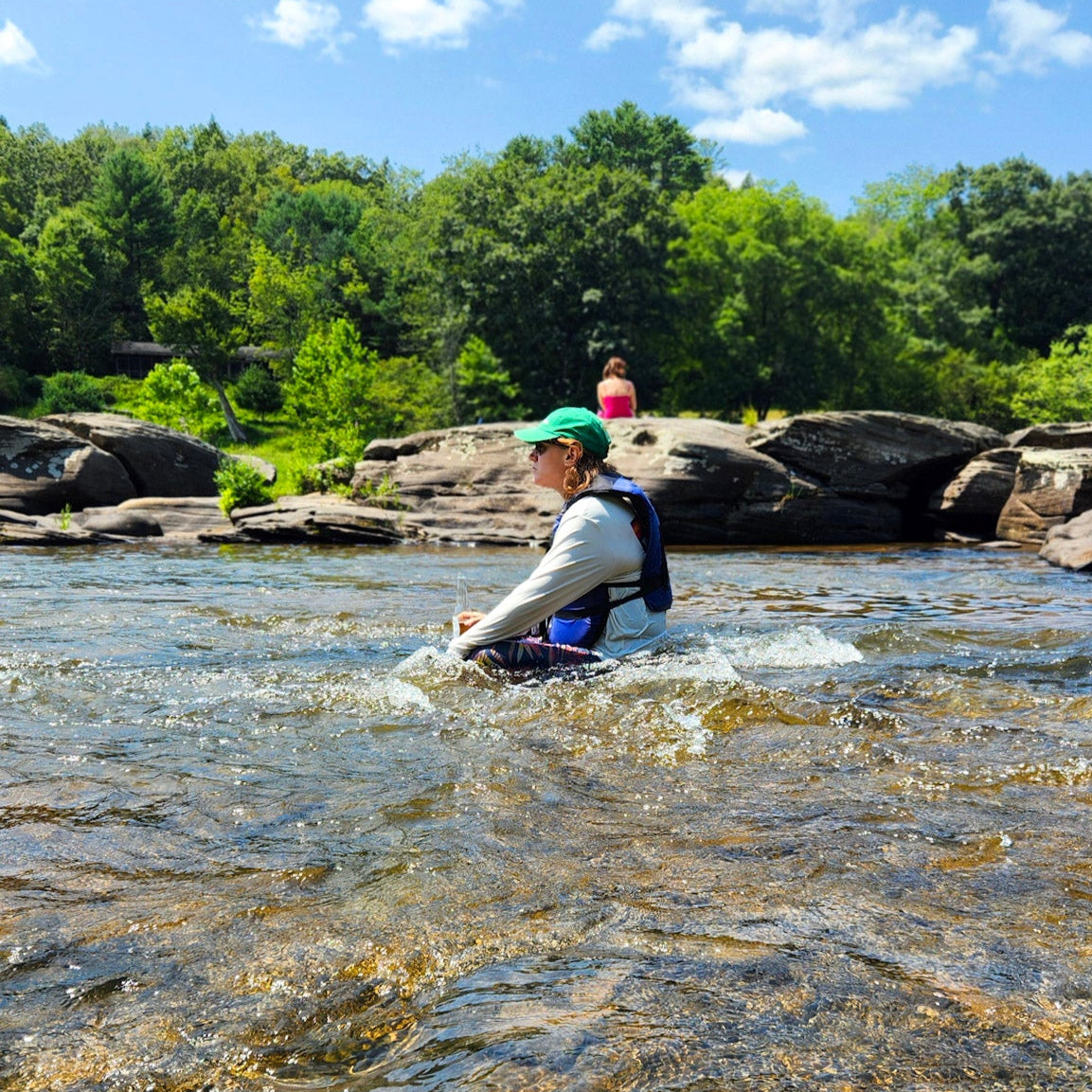 Person sitting at Skinner's Falls Rocky Beach with a life vest and trees in the background under a blue sky.