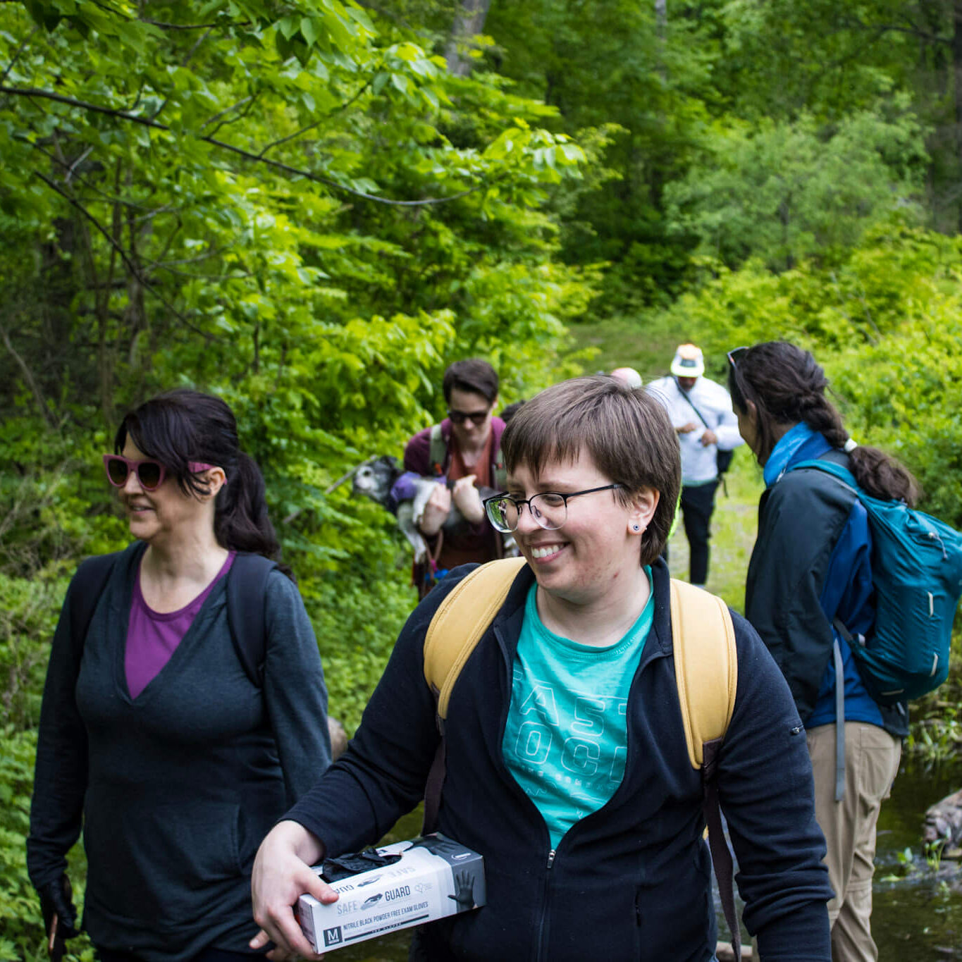 Group of people hiking in a forested area with a stream and wooden bridge in the background.