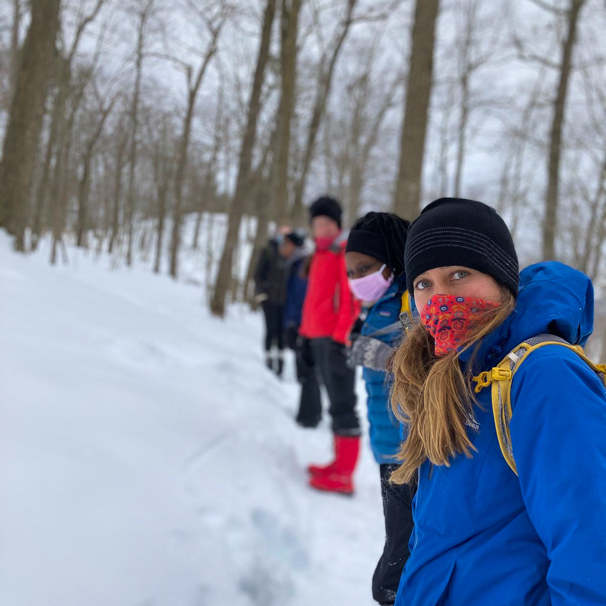 People walking in a snowy forest with one person in a blue jacket and colorful face mask.