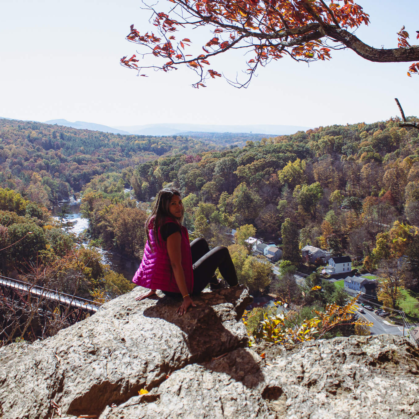 Person sitting on a rock overlooking a scenic valley with trees in autumn colors.