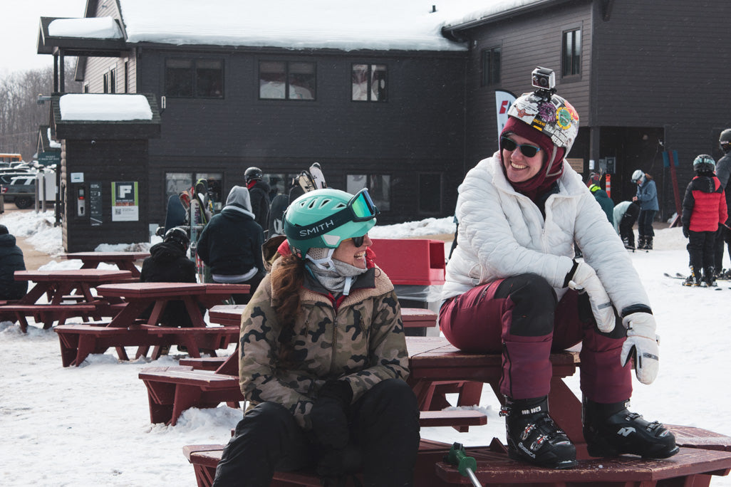 Two people sitting on a bench in a snowy outdoor setting with a building in the background.
