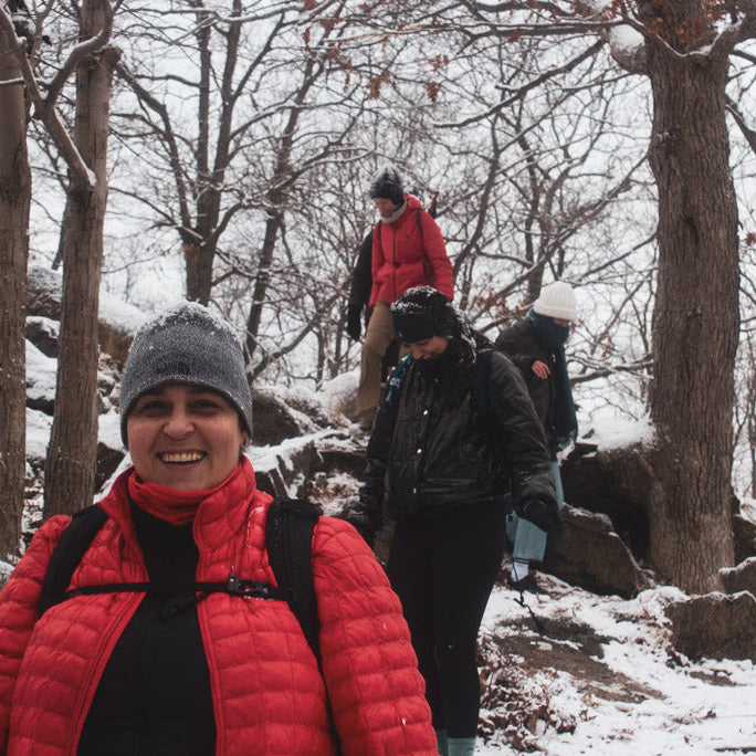 Person in a red jacket and gray beanie standing in a snowy forest with others in the background.