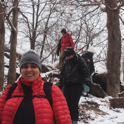 Person in a red jacket and gray beanie standing in a snowy forest with others in the background.